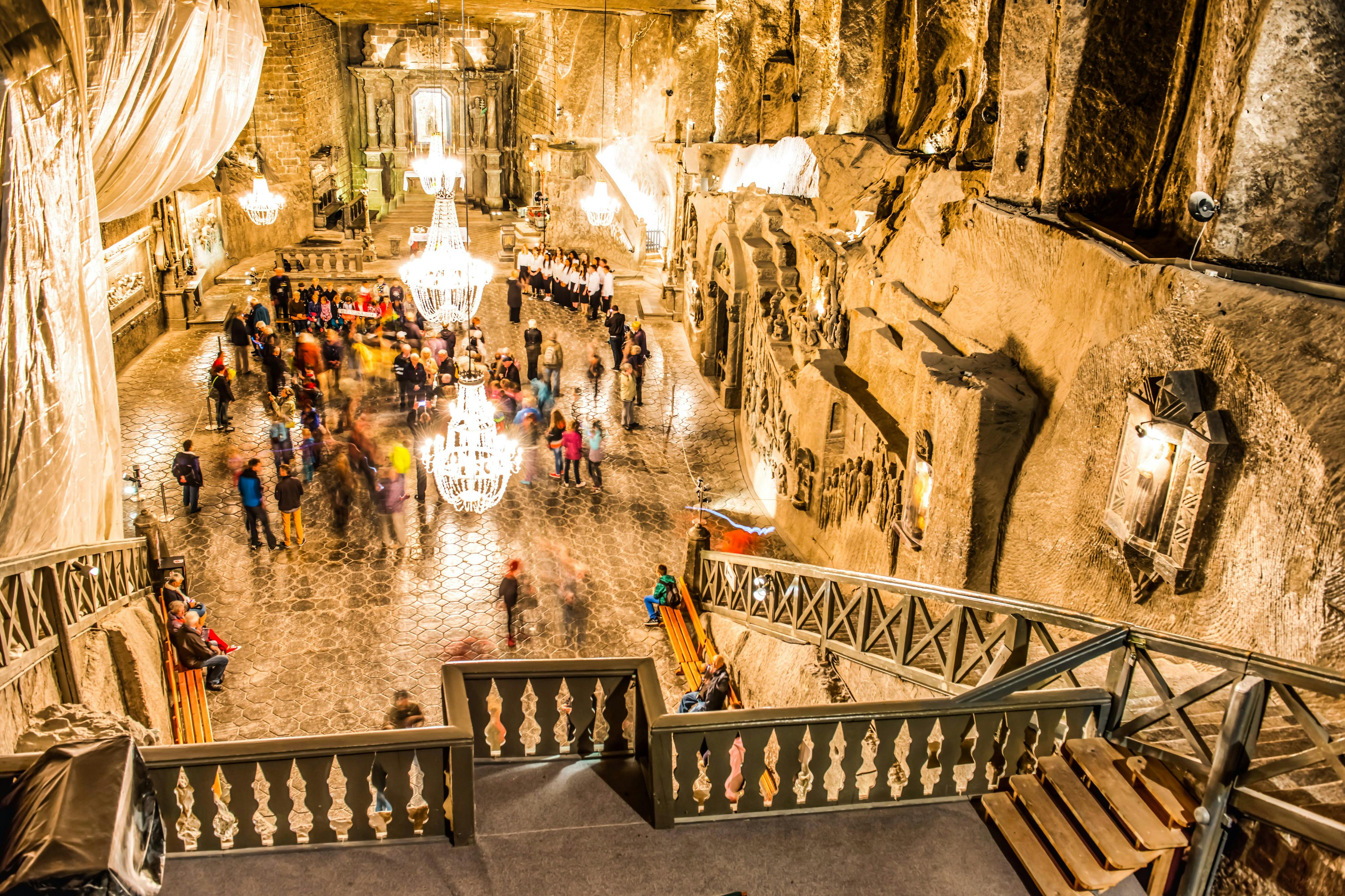 An ornate underground hall lit by chandeliers with a staircase, stone carvings on walls, and people walking around on a polished floor.