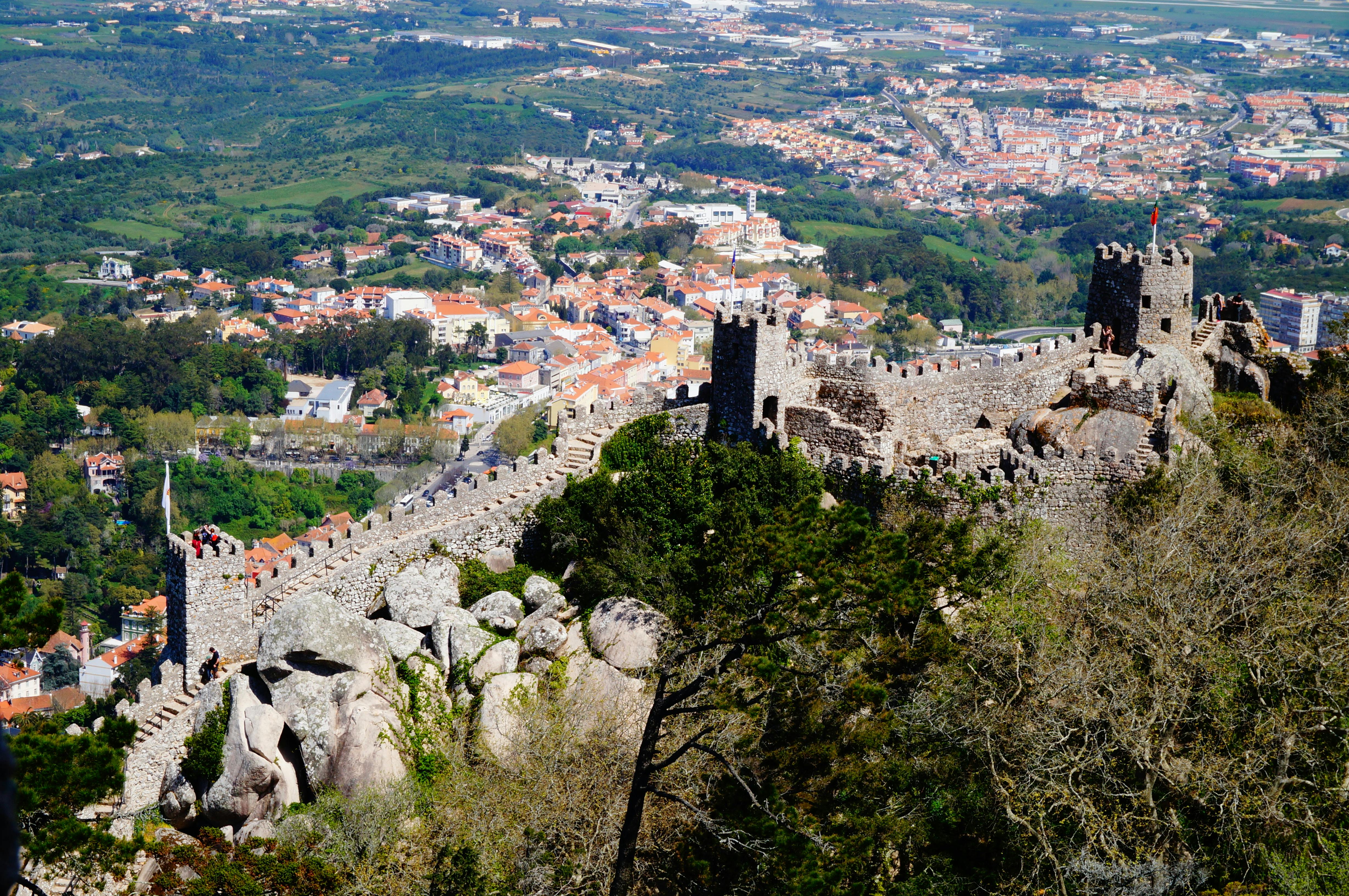 Древние каменные стены замка на вершине холма с видом на город и зеленый пейзаж внизу.