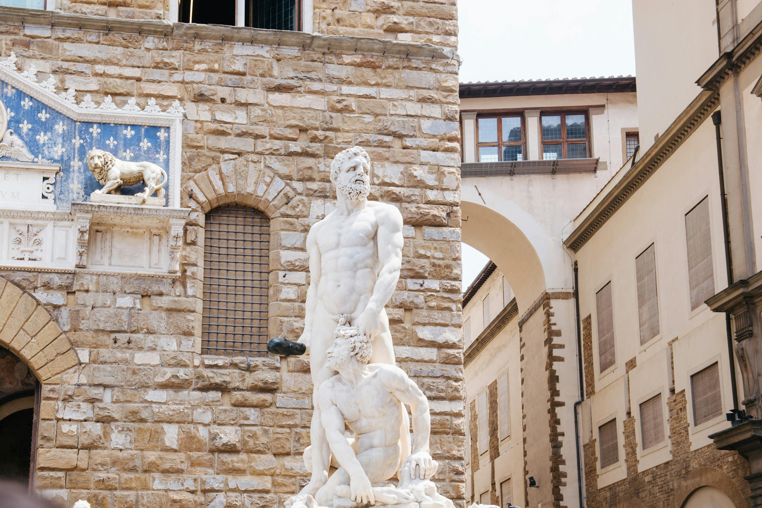 2人の筋肉質な男性の大理石の像。1人は立っていて、もう1人はひざまずいています。アーチ型の窓がある石造りの建物の上に立っています。