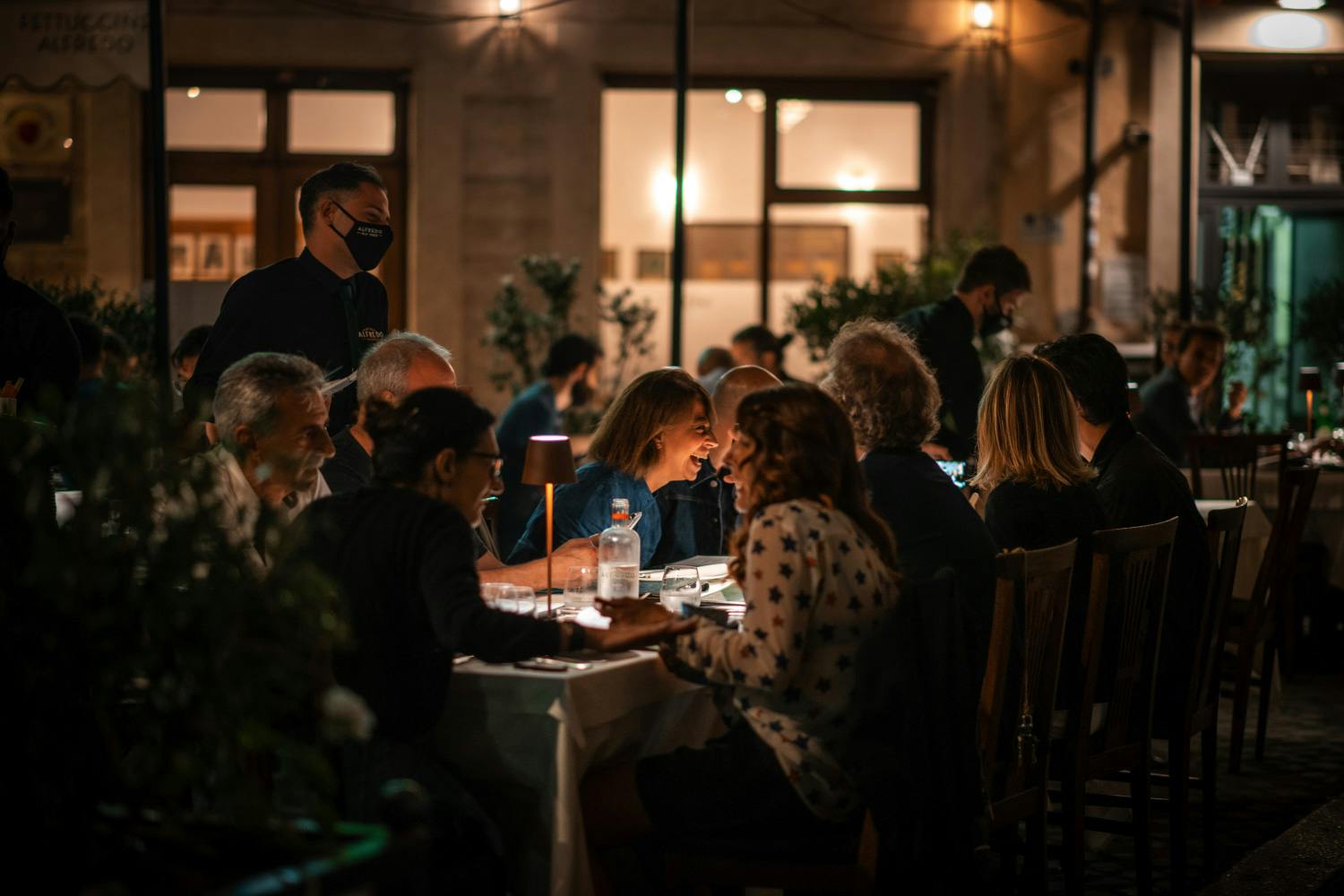 Люди ужинают на открытом воздухе ночью с сервером в маске, столиками, освещенными мягкими лампами, и теплым освещенным зданием на заднем плане.
