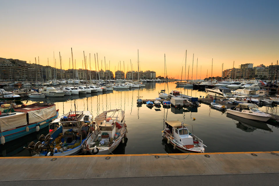 Boats docked in a marina at sunset, with buildings in the background and clear reflections on the calm water.