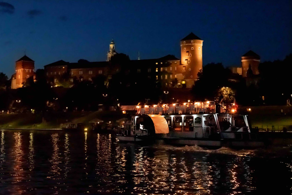 A boat illuminated with lights floats on a river at night; a historic building with towers is visible in the background.