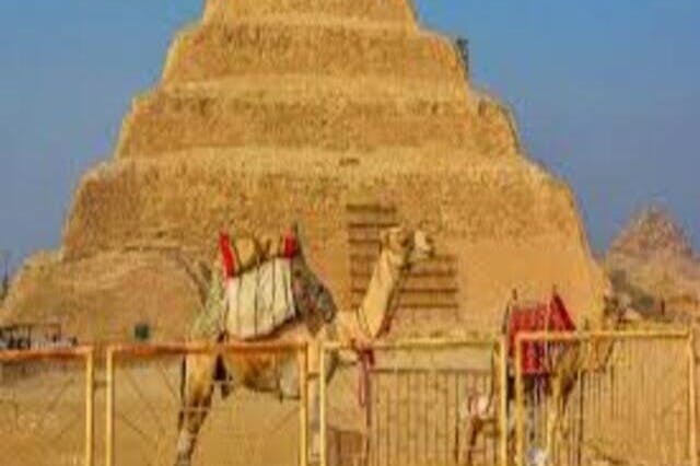 Ein Kamel steht vor einer Stufenpyramide unter strahlend blauem Himmel, wobei ein Zaun den Vorder- und Hintergrund trennt.