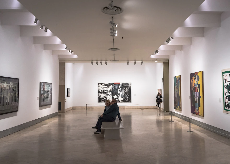 A person sits on a bench in a modern art gallery with various paintings on white walls, lit by ceiling spotlights.