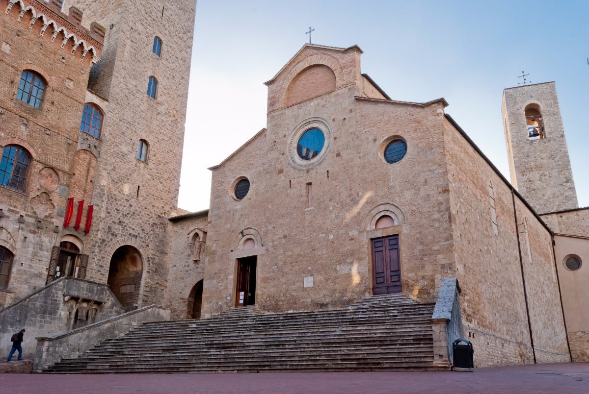 Kamienna fasada kościoła z drewnianymi drzwiami, okrągłymi witrażami, łukowym wejściem i dużą klatką schodową. Sąsiednia wysoka wieża.