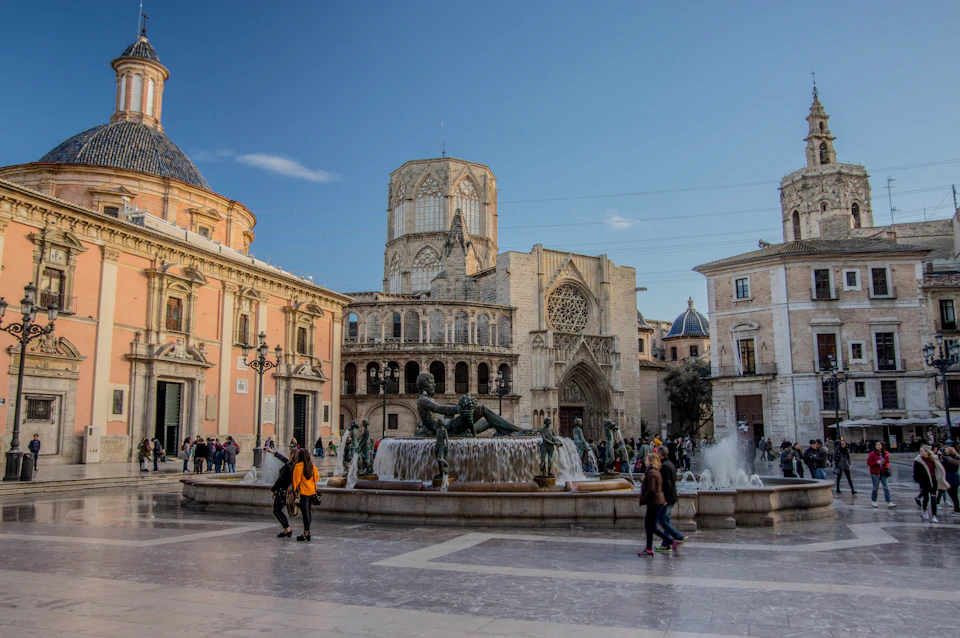 A bustling plaza with a large fountain, historic stone buildings, and people walking under a blue sky.