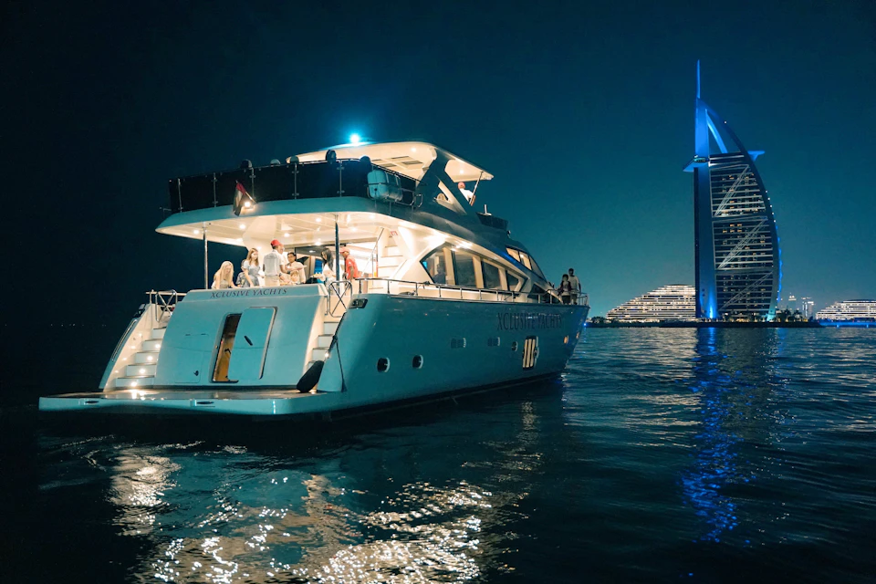 Luxurious yacht illuminated at night with people on deck, floating on water; a lit-up modern building stands in the background.