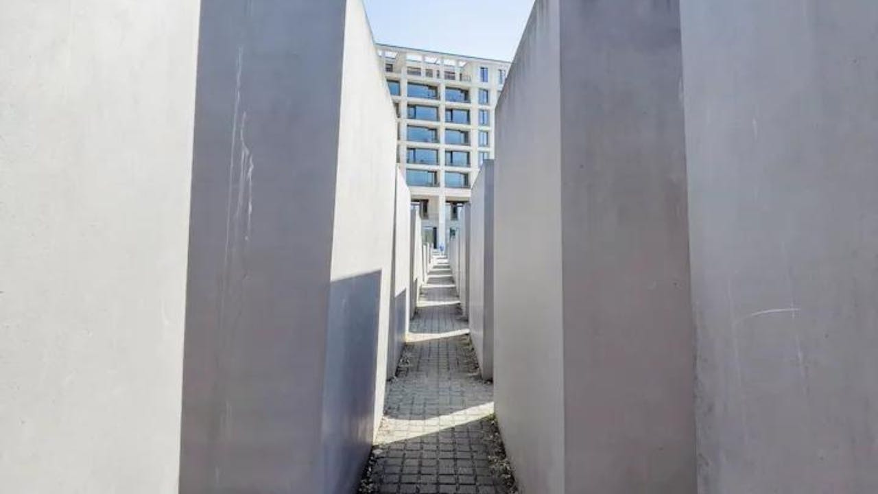 K moderní prosklené budově v pozadí vede úzký chodník lemovaný vysokými betonovými bloky.