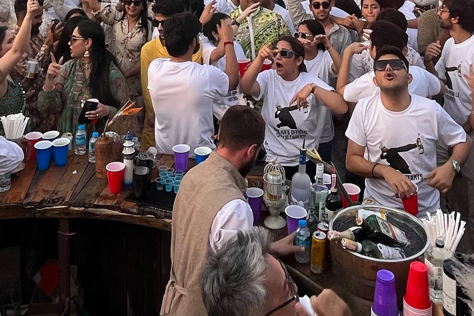 A lively gathering on a riverboat with people wearing white shirts, enjoying a party near a cityscape and a bridge.
