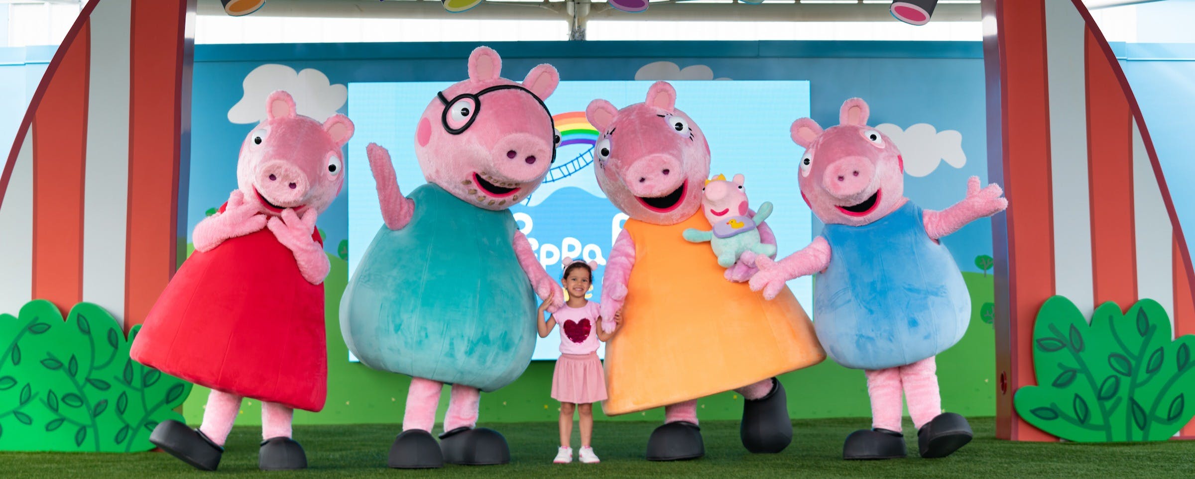 明るい背景のステージに、カラフルな衣装を着た漫画のブタのキャラクターのマスコットを4つ持った子供が立っています。