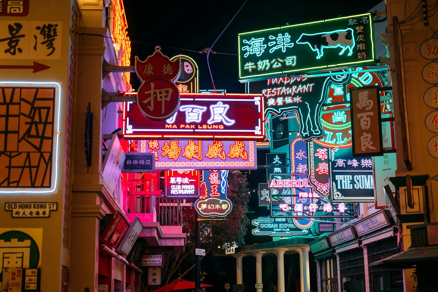 A colorful street scene at night, featuring vibrant neon signs in various languages and designs above storefronts.