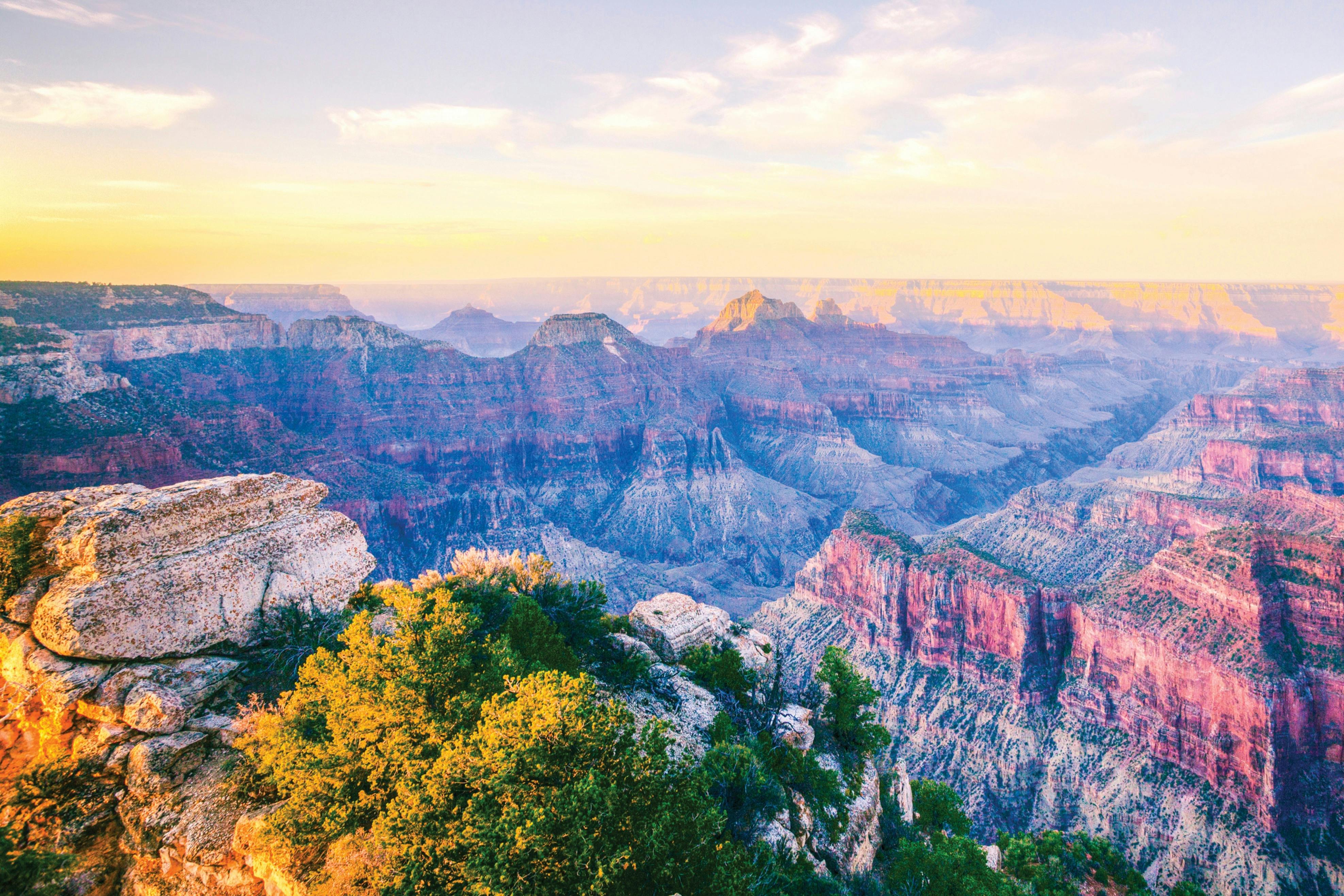 A scenic view of the Grand Canyon with colorful rock formations, cliffs, and vegetation under a partly cloudy sky.