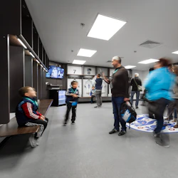 En grupp människor, inklusive barn, i ett omklädningsrum för sport med tröjor som hänger i bås och en TV som visar bilder.