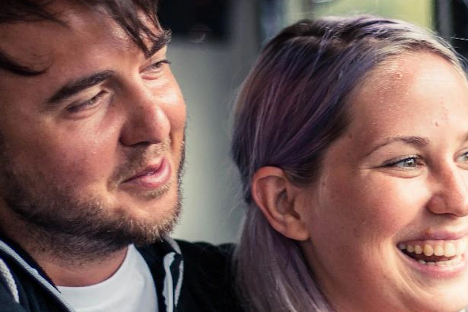 A man and woman embrace and smile inside a bus, with windows and a dimly lit interior visible in the background.
