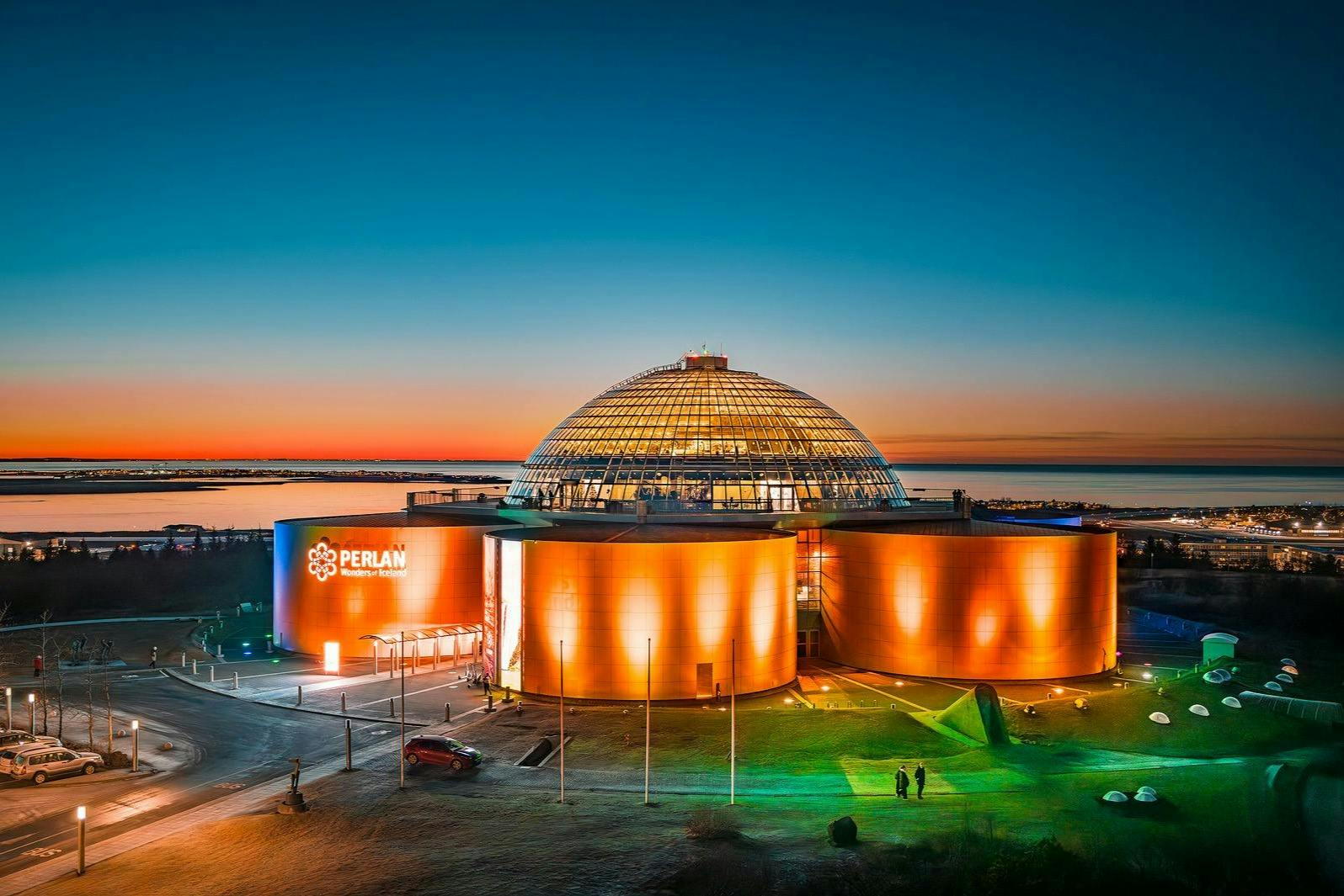 En stor bygning med en glaskuppel og oplyste orange vægge, mærket 'Perlan', ved solnedgang med udsigt over kysten.