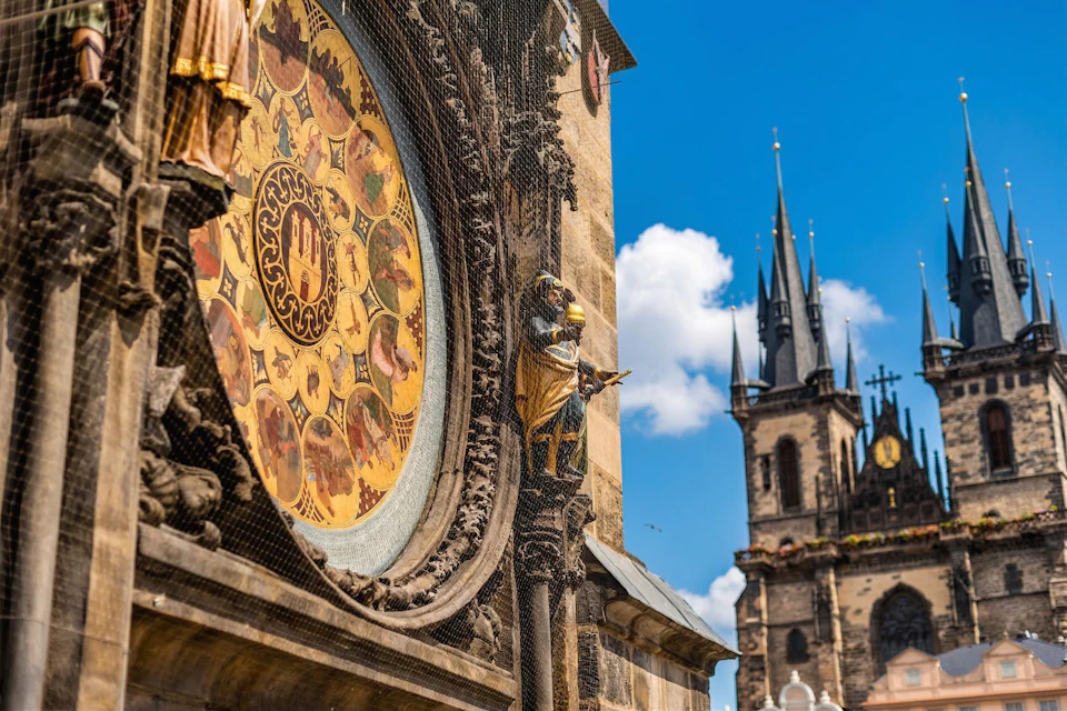 A close-up of a decorative astronomical clock in Prague, with a church spire and blue sky in the background.