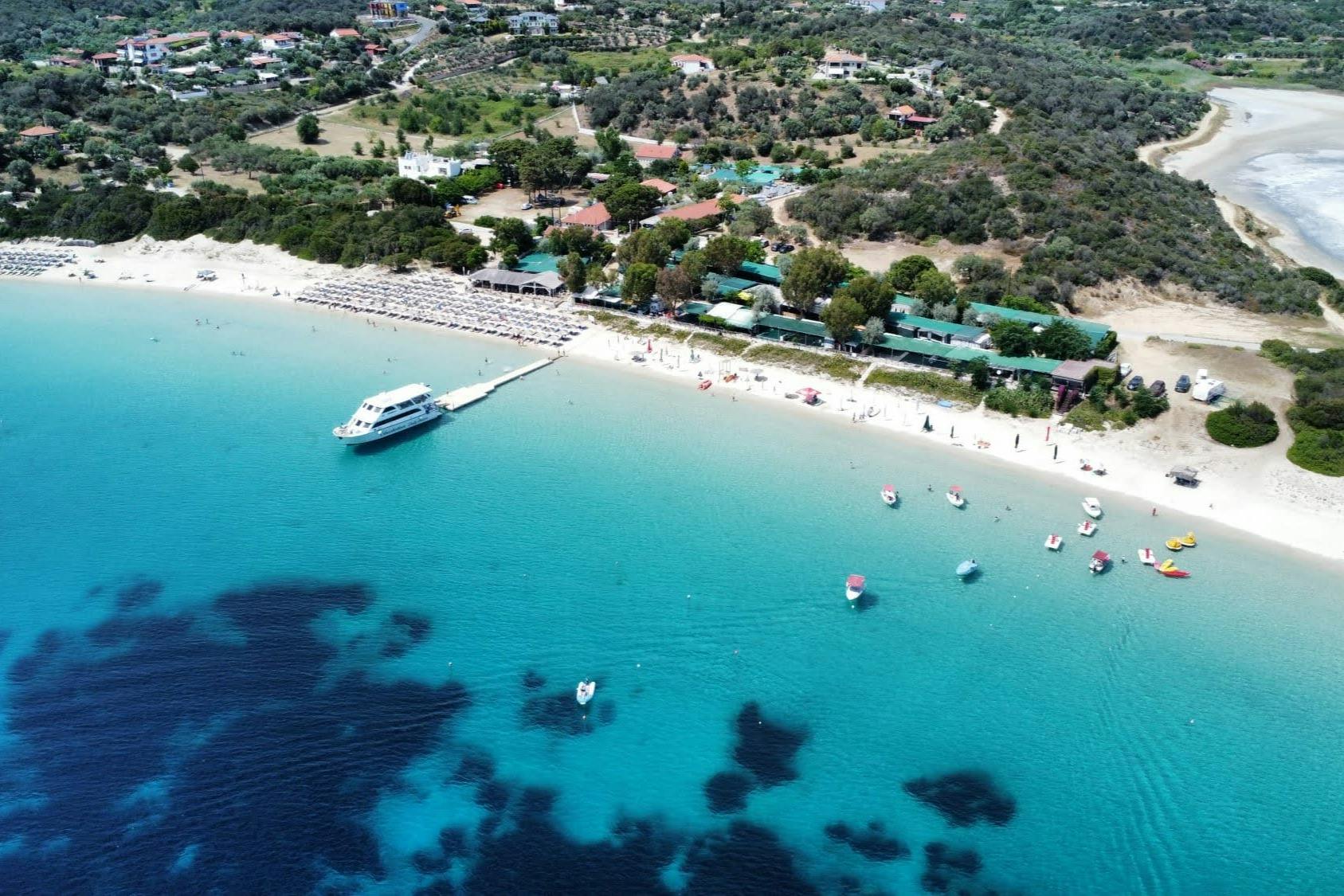 Luchtfoto van een strand met turquoise water, een witte boot op een pier en gebouwen omgeven door groen op de achtergrond.