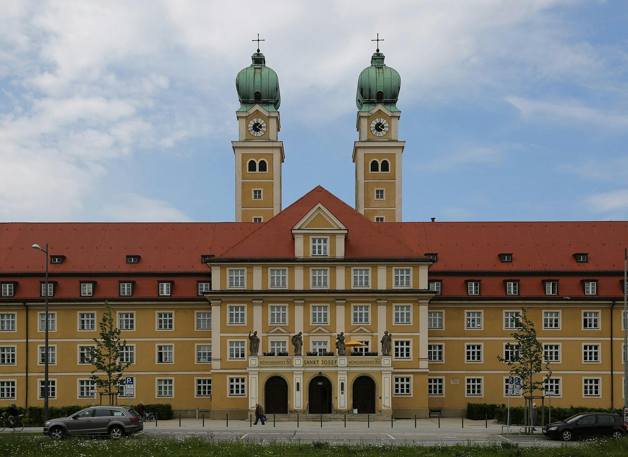 赤い屋根と2つの時計塔が緑色のドームで覆われた大きな黄色い建物。 前方には車と人が見えます。