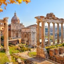 Ancient Roman ruins with tall columns and a grand domed building under a clear blue sky, with yellow leaves in the foreground.