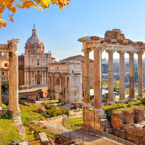 Ancient Roman ruins with tall columns and a grand domed building under a clear blue sky, with yellow leaves in the foreground.