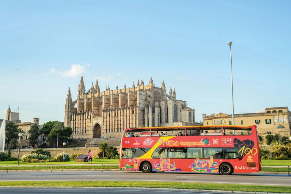 A red double-decker sightseeing bus drives past a large, gothic cathedral with ornate spires under a clear blue sky.