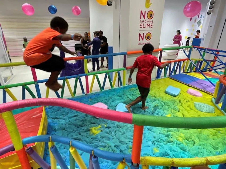 Children playing on a colorful indoor obstacle course with soft, padded surfaces and signs prohibiting slime and running.