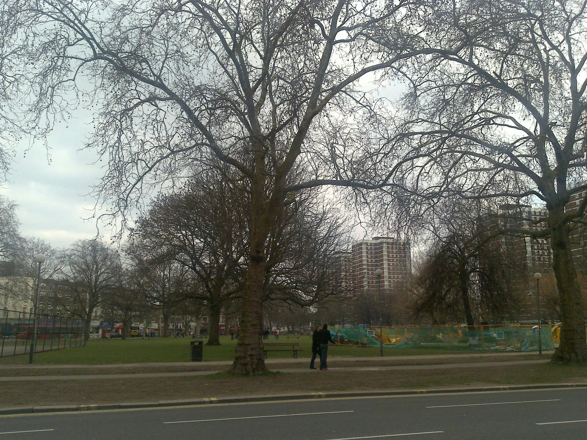 Park z drzewami bezlistnymi, teren budowy ogrodzony zieloną siatką, kilka osób spacerujących, a w tle wysokie budynki.