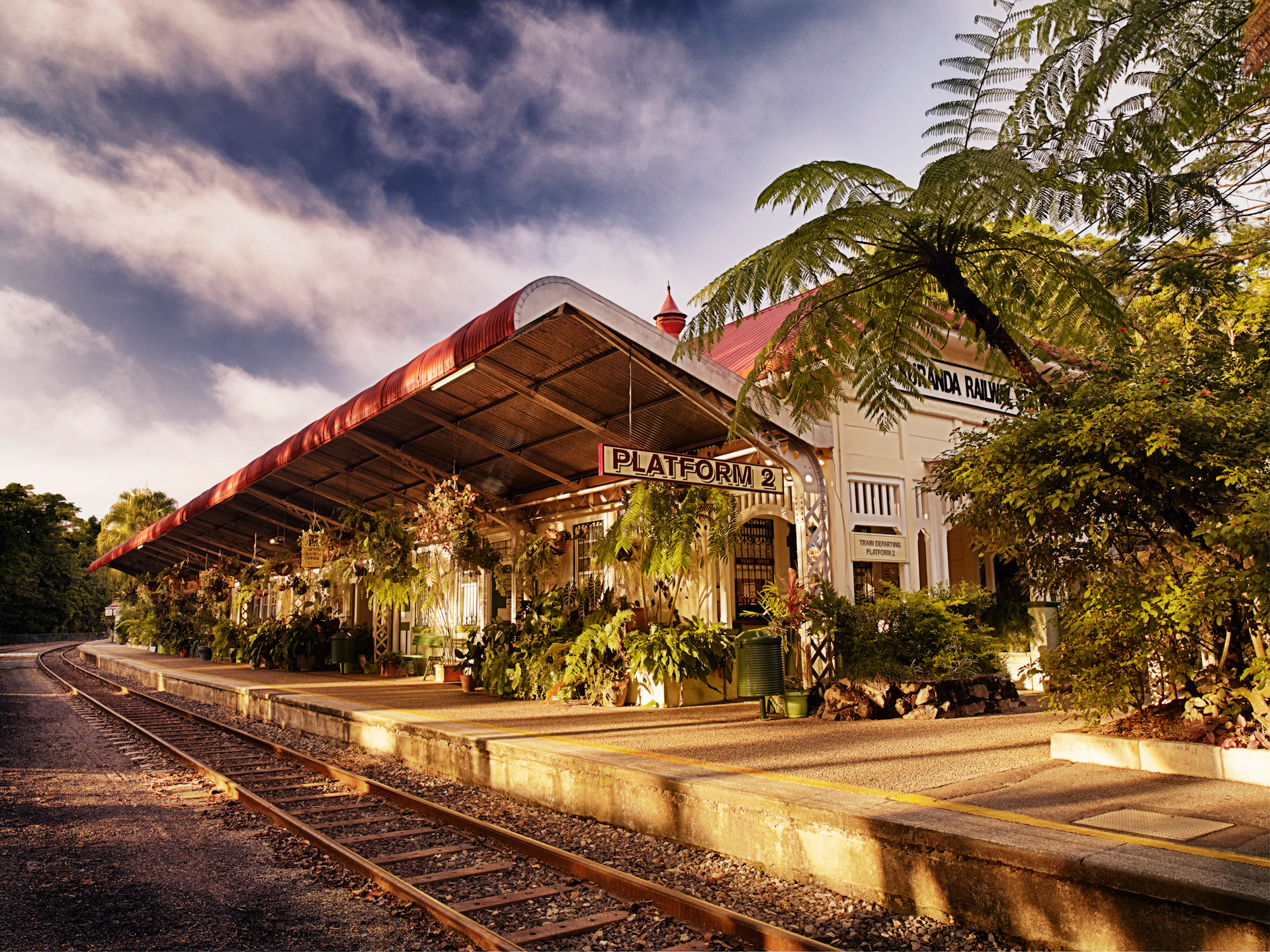 Outdoor train platform with plants, a sign reading "PLATFORM 2," and a red-roofed structure under a cloudy sky.