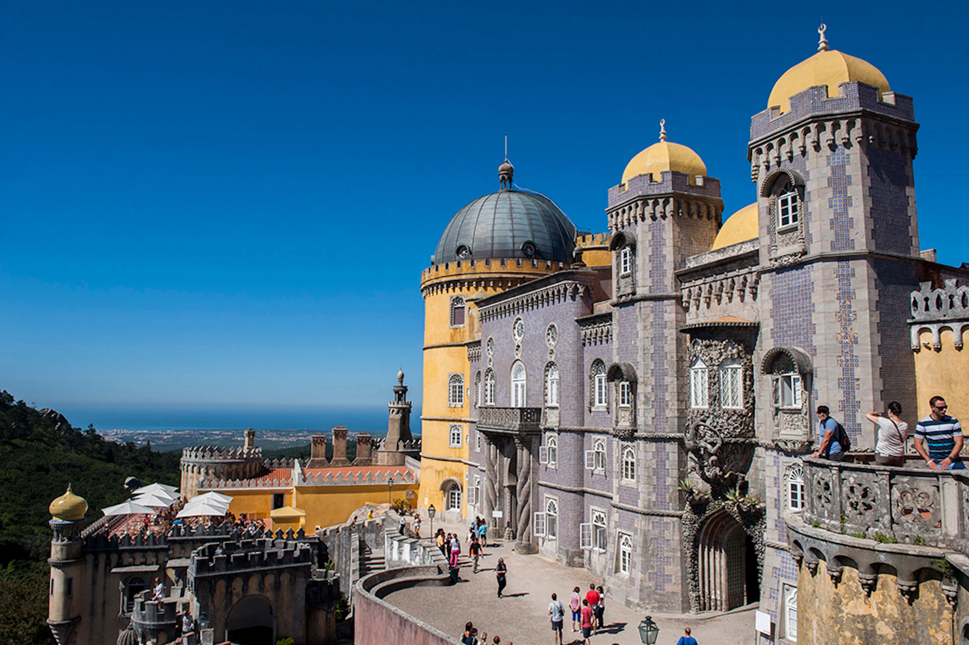 People walking outside a colorful, ornate palace with yellow and purple towers under a clear blue sky.