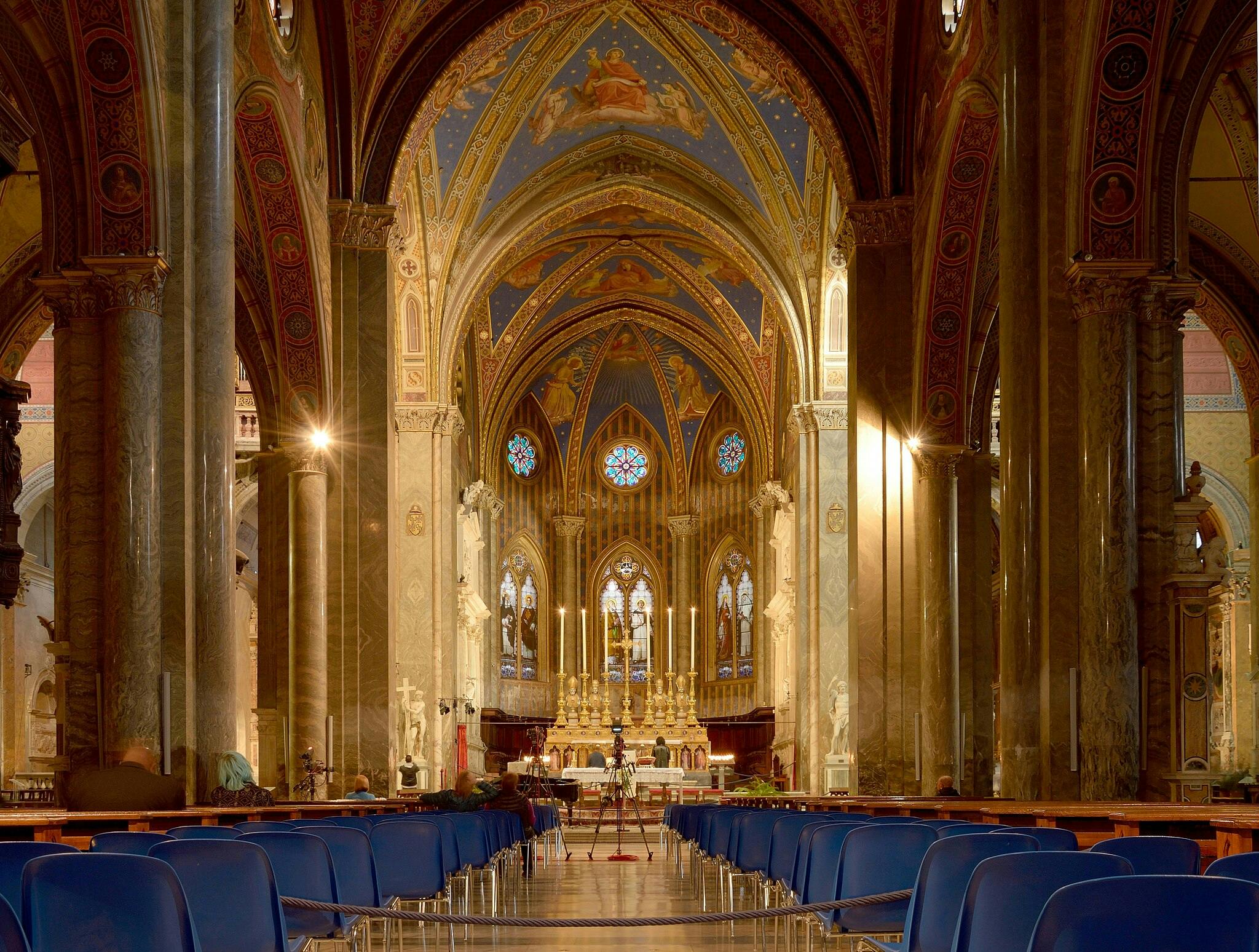Ornate cathedral interior with vaulted ceilings, stained glass windows, rows of chairs, and an elaborate altar at the end.