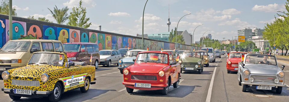 A convoy of colorful classic cars drives past a graffiti-covered wall in an urban area on a sunny day, with a TV tower in the background.