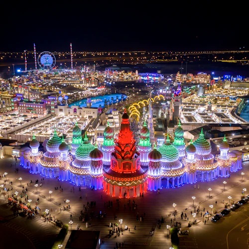 Aerial view of an illuminated cultural village at night featuring colorful domes and towers, with people walking and a Ferris wheel in the background.