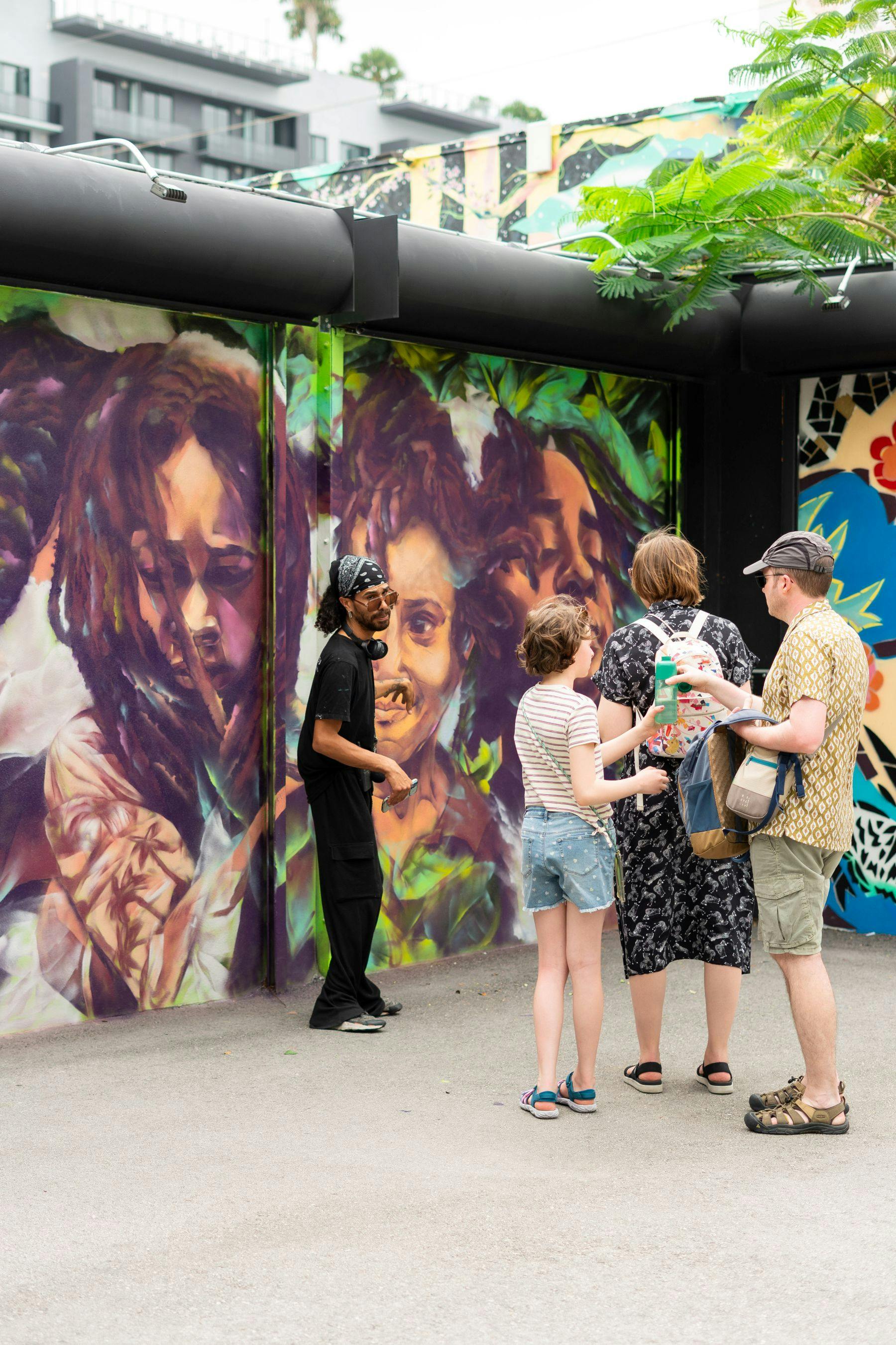 人们欣赏充满活力的壁画艺术，描绘面孔和树叶，艺术家在场。 场景包括郁郁葱葱的绿色植物。