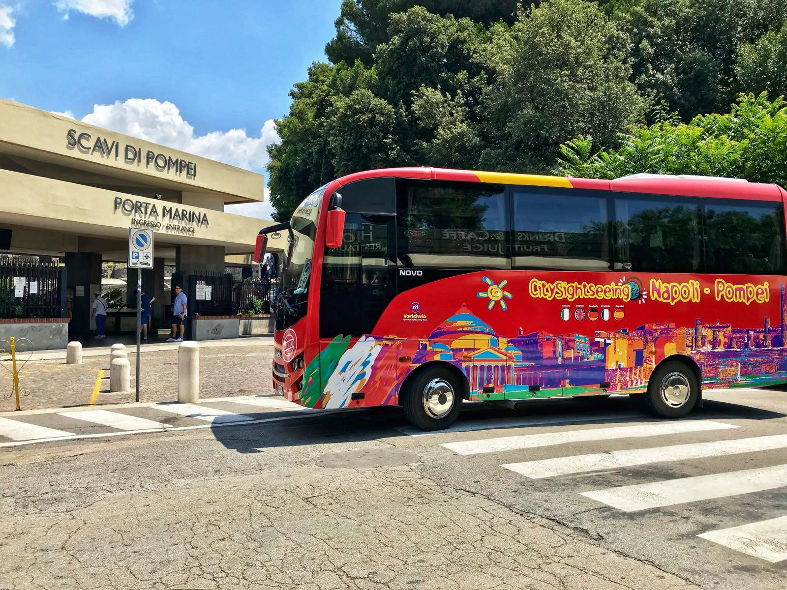 Ένα πολύχρωμο λεωφορείο για τα αξιοθέατα είναι σταθμευμένο κοντά στην είσοδο της Porta Marina των ερειπίων της Πομπηίας, με δέντρα και ένα γαλάζιο ουρανό στο παρασκήνιο.