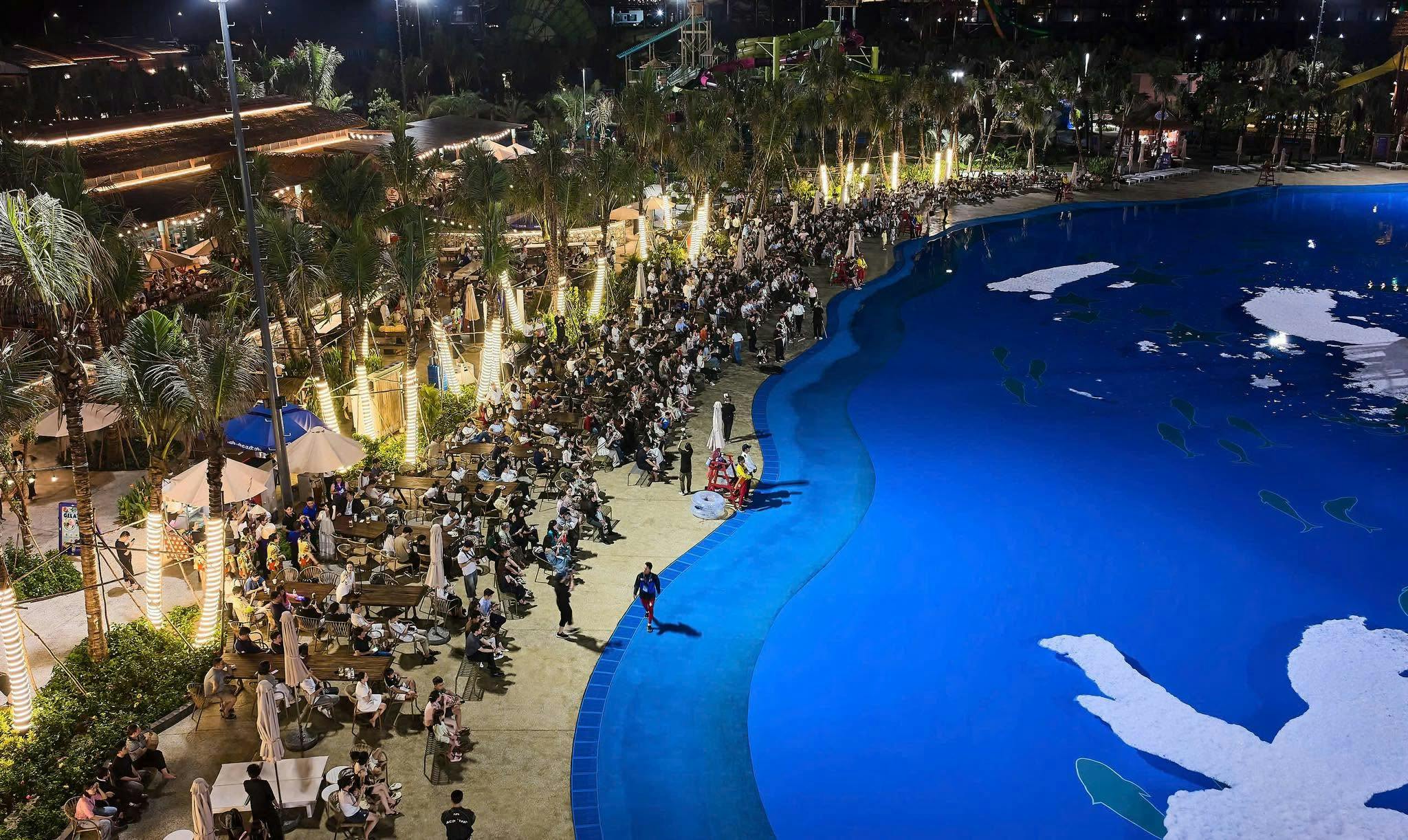 A large gathering of people seated and walking by a brightly lit blue artificial pool at night, surrounded by palm trees.