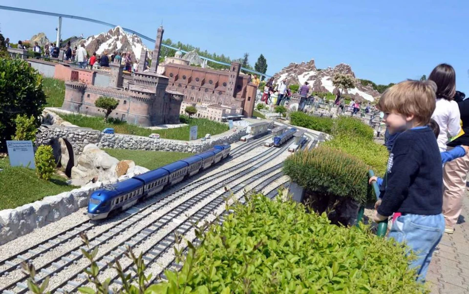 Miniature trains on tracks in a detailed scale model of a town, surrounded by greenery. People are observing the display.