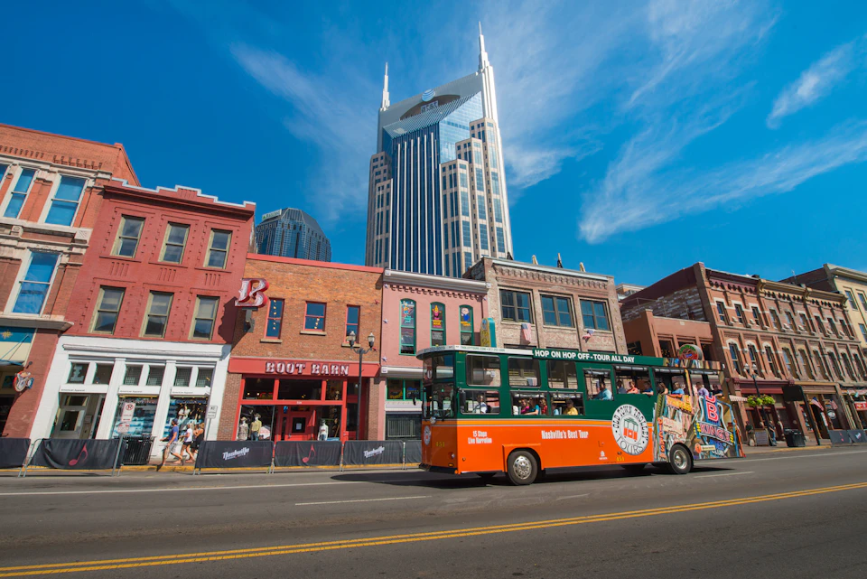 A lively street with a green and orange tour bus passing by colorful buildings, including a bar, under a clear blue sky.