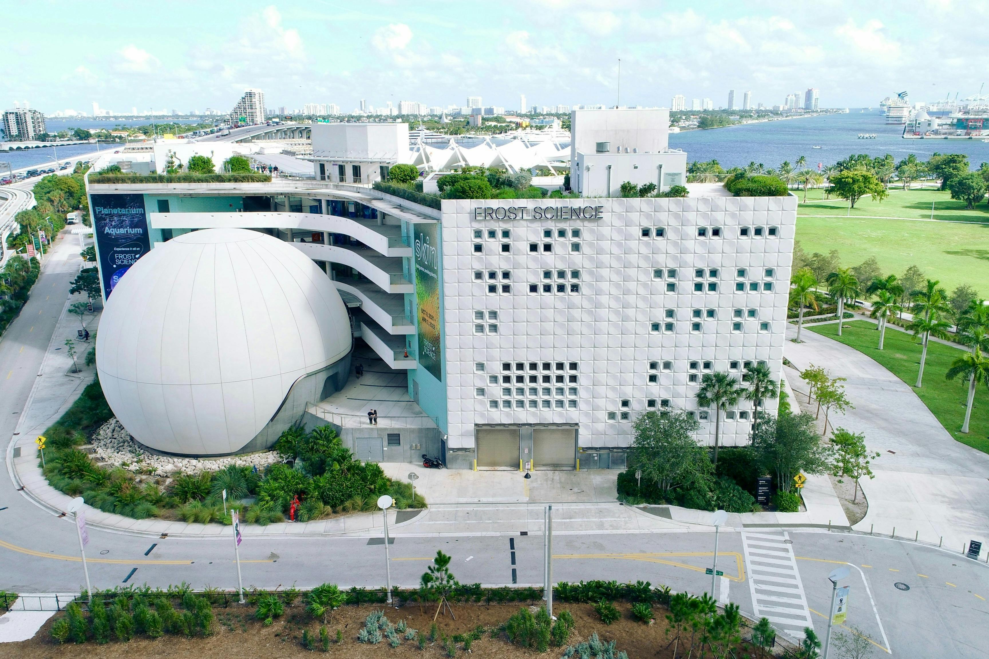 Luftfoto af en moderne videnskabsmuseumsbygning med en kuppel, urbane omgivelser og en blå himmelbaggrund.
