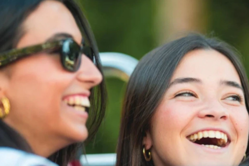 Two women smiling and laughing outdoors, one wearing sunglasses, with a blurred green background.