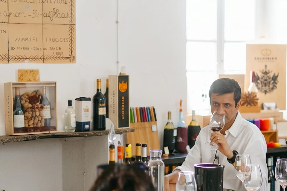 A group of people seated around a table with wine glasses, while one person stands and speaks in a cozy room.