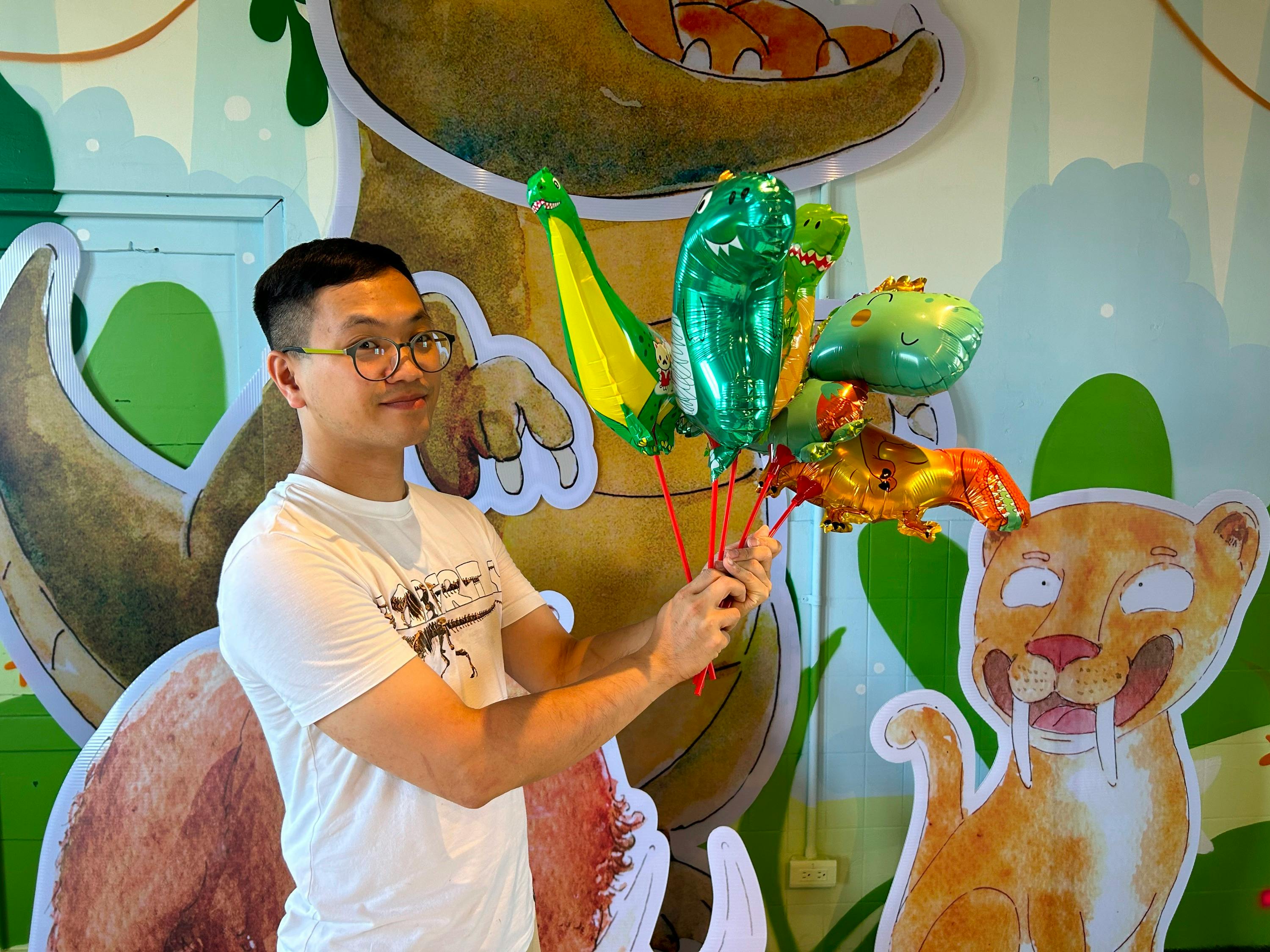 A person holding colorful dinosaur-shaped balloons in front of a cartoon dinosaur backdrop, wearing glasses and a white shirt.