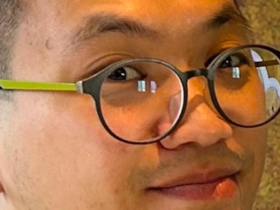 A person holding colorful dinosaur-shaped balloons in front of a cartoon dinosaur backdrop, wearing glasses and a white shirt.