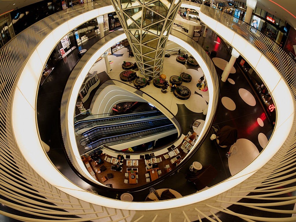 A multi-level shopping mall with lit circular walkways, escalators, seating areas, and various storefronts.