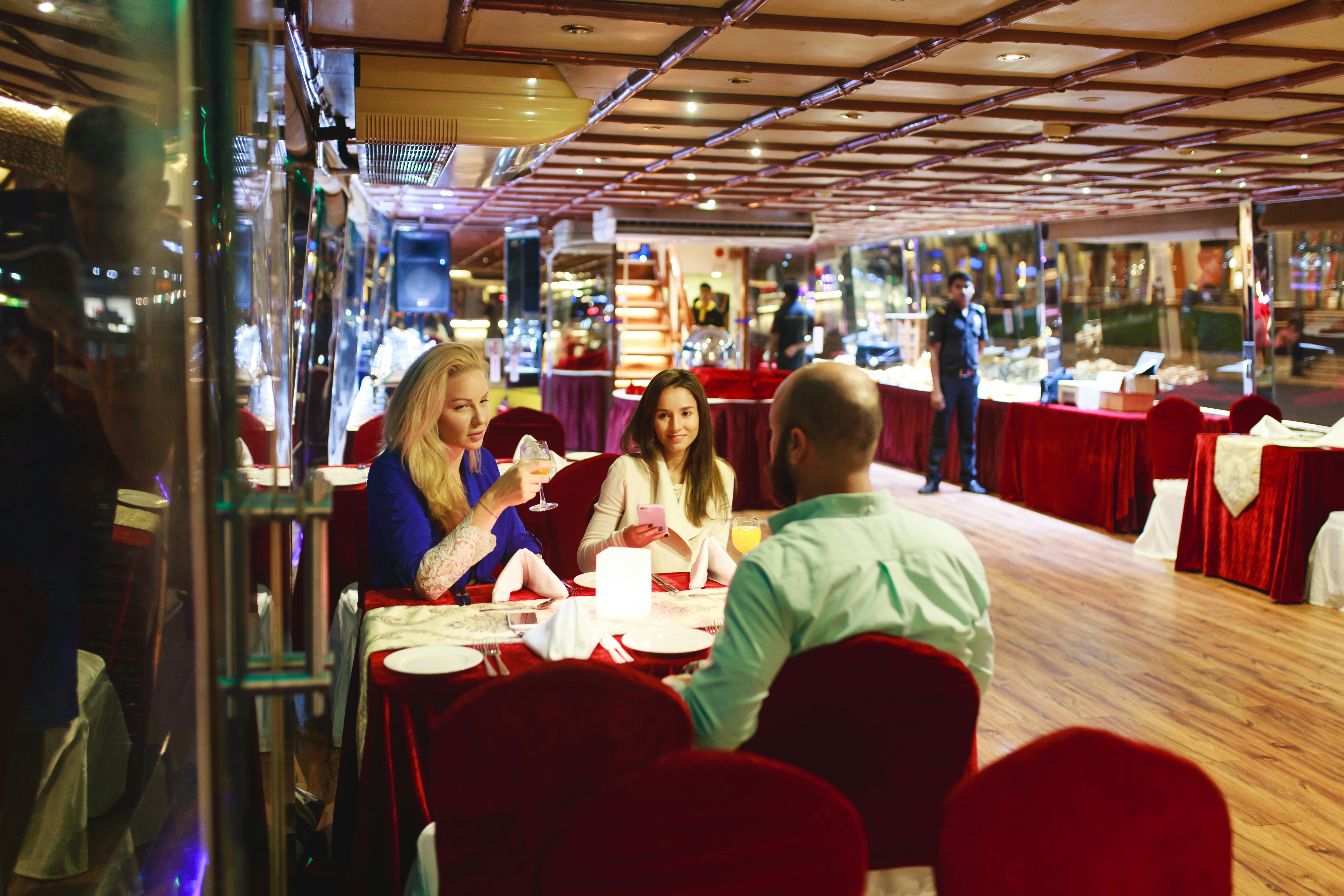 Three people seated at a restaurant table in a well-lit, upscale dining area, engaged in conversation, with a server in the background.