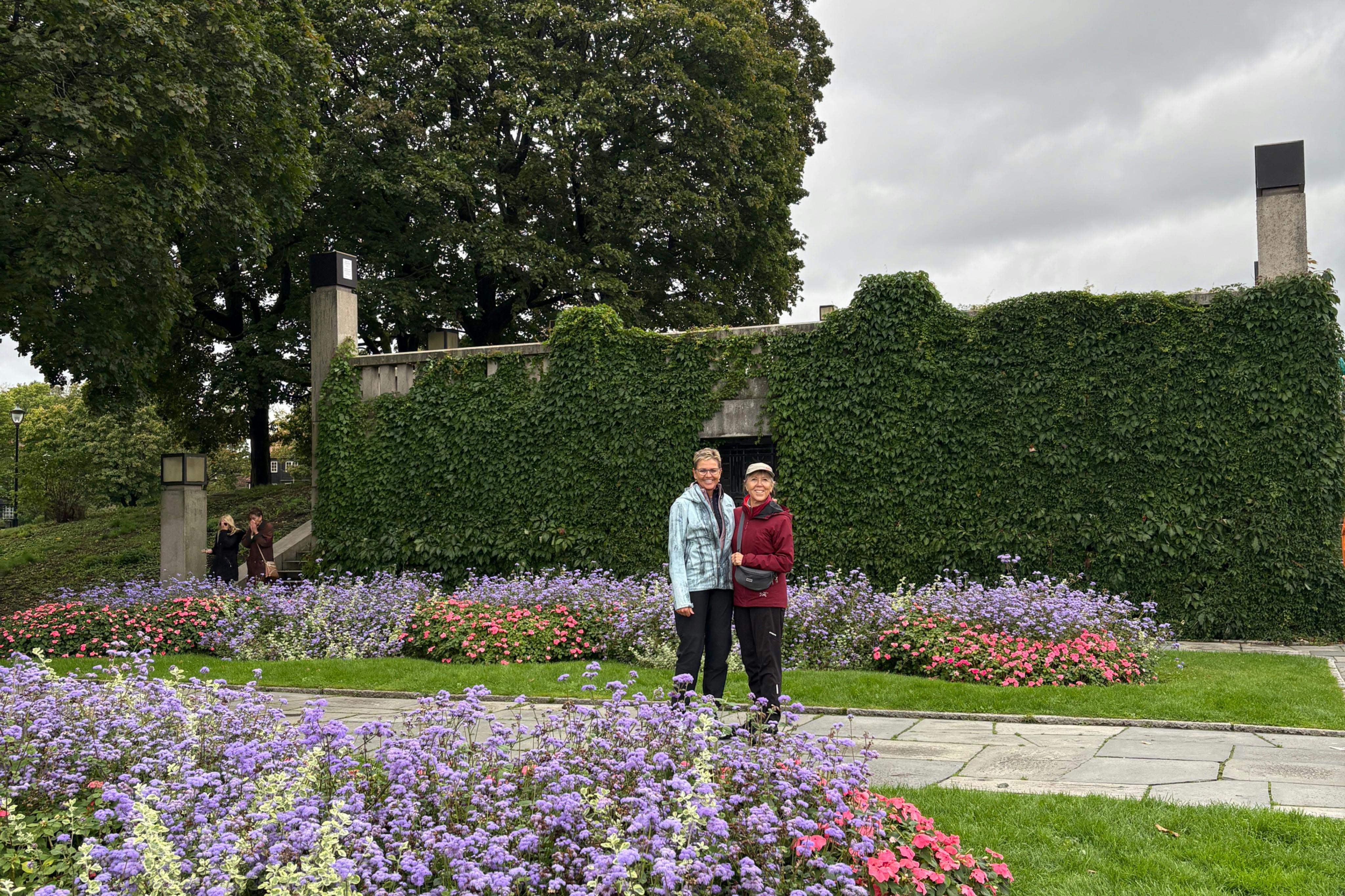 两个人站在一个紫色和粉色花朵的花园里的小径上，前面的墙上覆盖着绿色的常春藤。多云的天空。