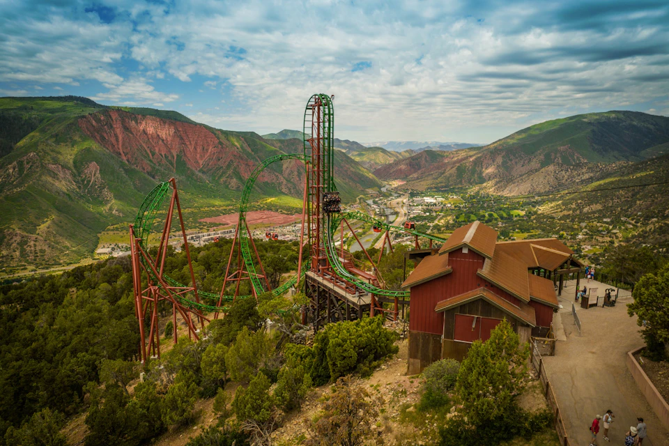 Green rollercoaster in a mountainous landscape with red rock formations, adjacent to brown buildings amidst trees under a cloudy sky.