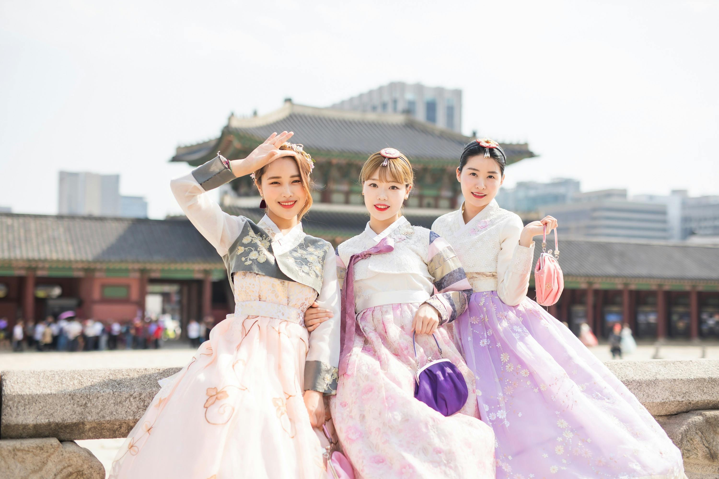 Three women wearing traditional Korean hanbok dresses pose happily in front of a historic building on a sunny day.