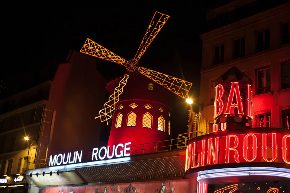 Night view of the illuminated Moulin Rouge, showing a red windmill with lights and surrounding neon signs.