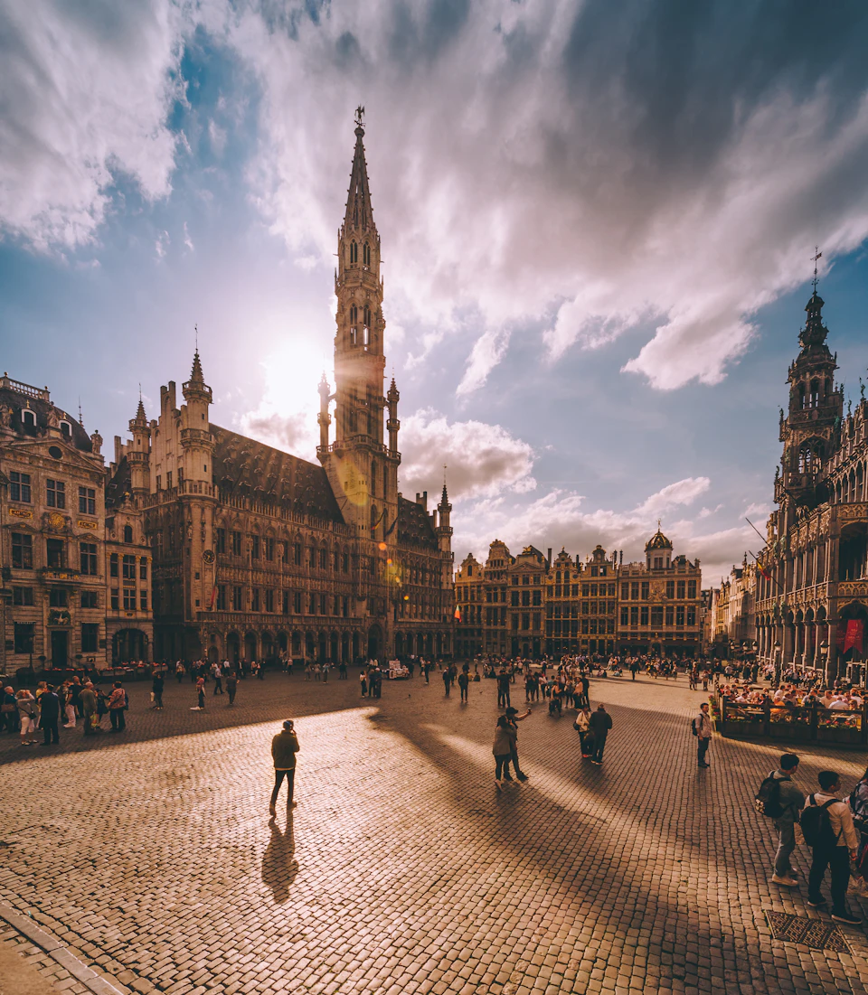 Sunlit square with historic buildings, a tall central tower, and people walking and gathering. Sky is partly cloudy.