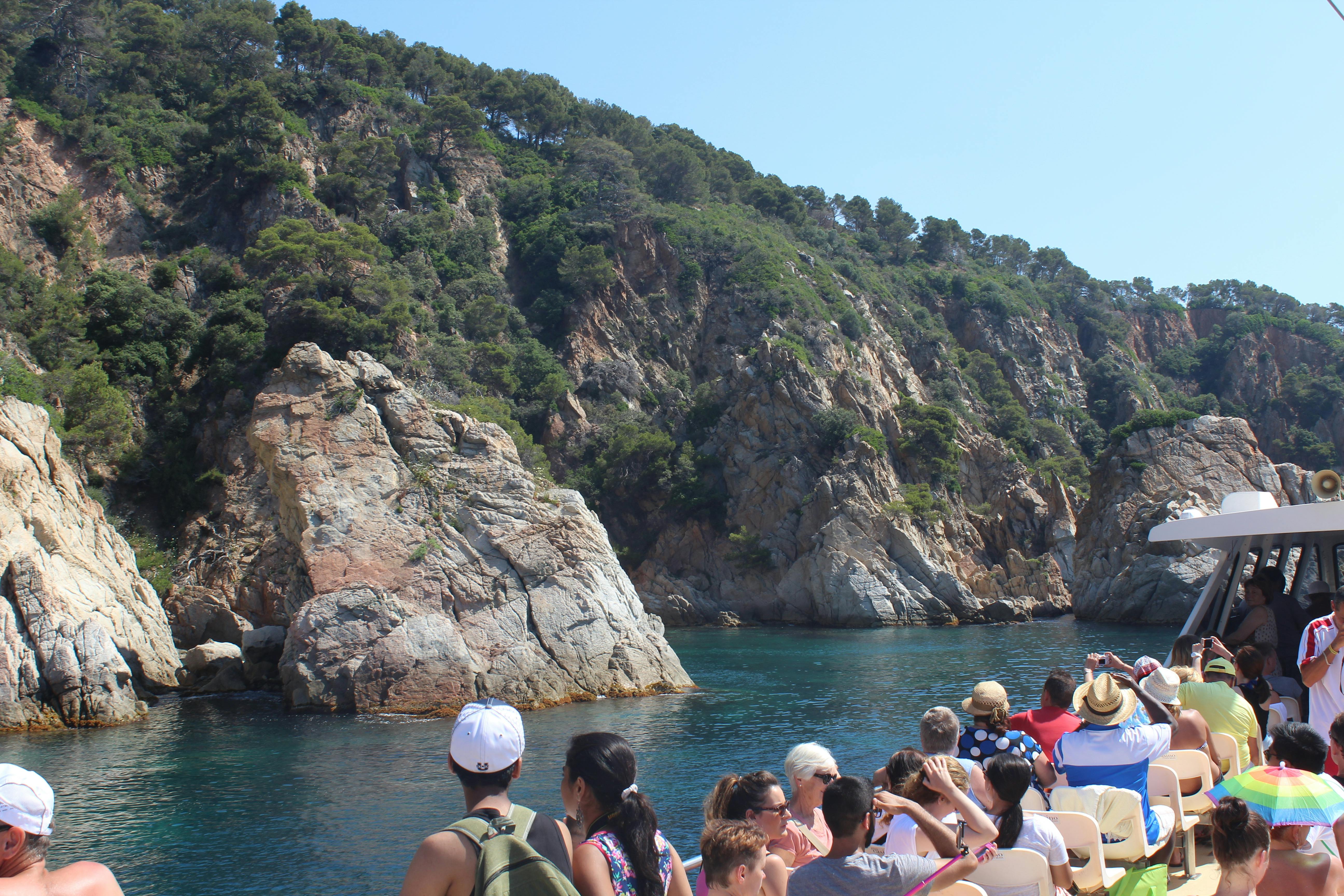 在岩石海岸线附近的船上观光的游客，在明亮的天空下有郁郁葱葱的绿树和清澈的蓝色水域。